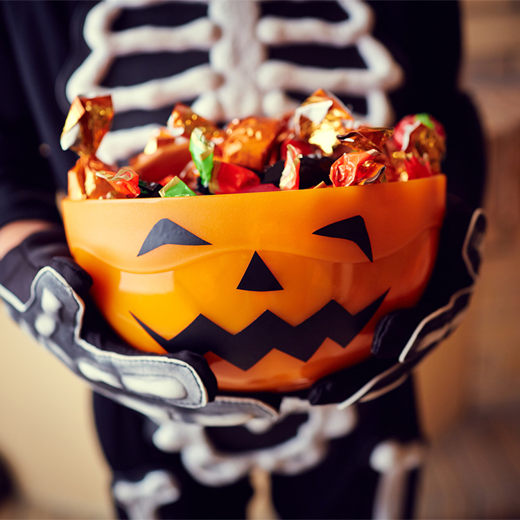 Kid in skeleton costume holds jack o'lantern candy bowl full of generic candies