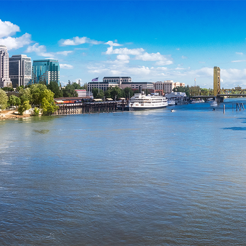View of downtown Sacramento from the river. Office buildings stand on the right, a bridge on the left, and in the foreground is the river. A docked boat can be seen on the downtown side of the river.