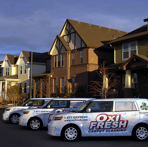 Three Oxi Fresh vans sit outside a row of suburban homes. Light from the sunrise highlights the cars and the buildings.