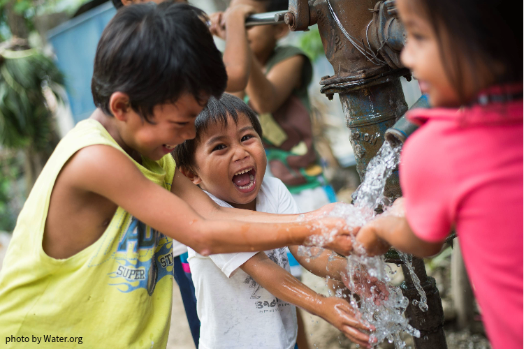 This image shows a group of young children laughing as they play in the water flowing from a tap.