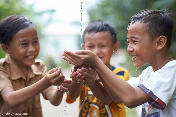 Three smiling, laughing children cup their hands together to catch a stream of water.