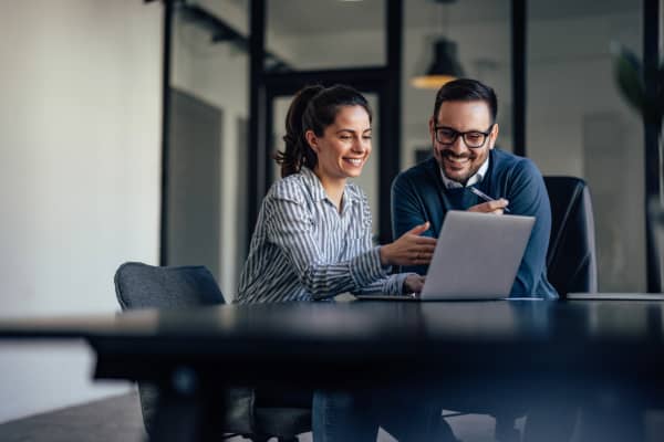 Two professionals smile while talking about what they're seeing on a laptop in an office.