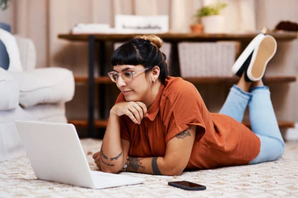 A woman lays on her carpet while looking at her laptop.
