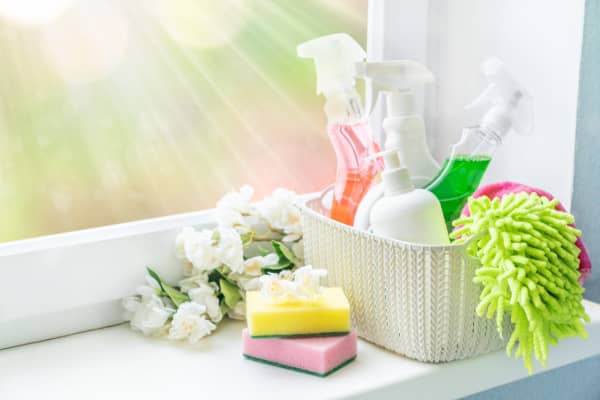 A basket of cleaning items and flowers sitting on a white windowsill.