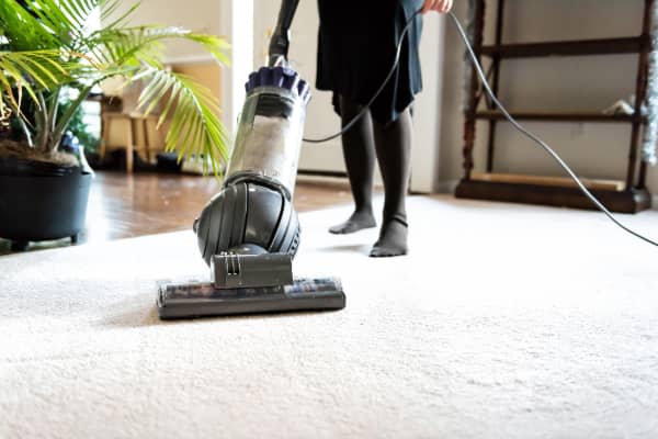 A woman vacuums her white carpet while holding the vacuum wire suspended in her left hand.